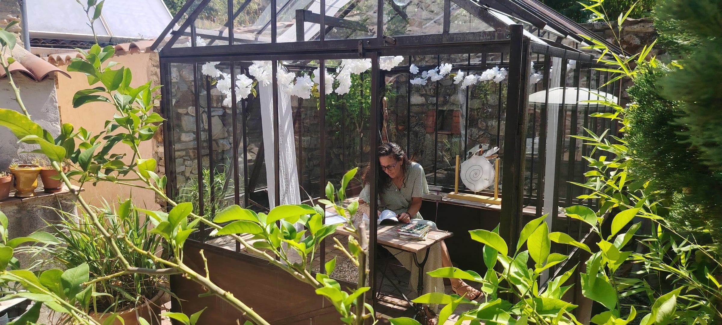 Femme assise à une table en bois, écrivant dans un carnet à l'intérieur d'une véranda vitrée entourée de plantes vertes et de guirlandes de fleurs blanches
