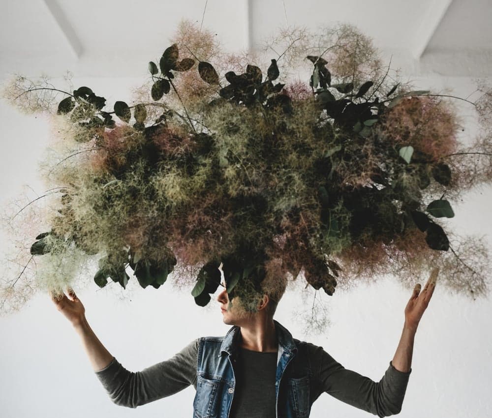 Personne en veste en jean, bras levés, le visage disparaissant sous une immense composition suspendue de feuillages et de fleurs vaporeuses