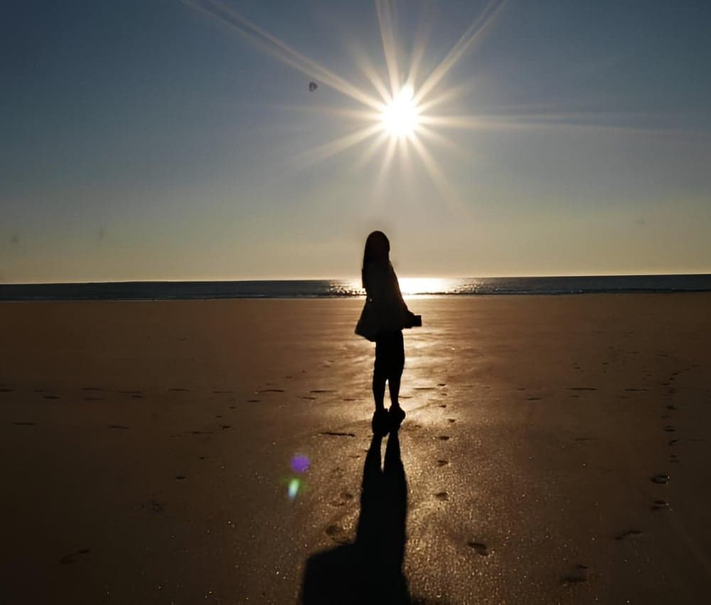 Silhouette d'une femme debout sur une plage face à la mer, sous un soleil éclatant en contre-jour